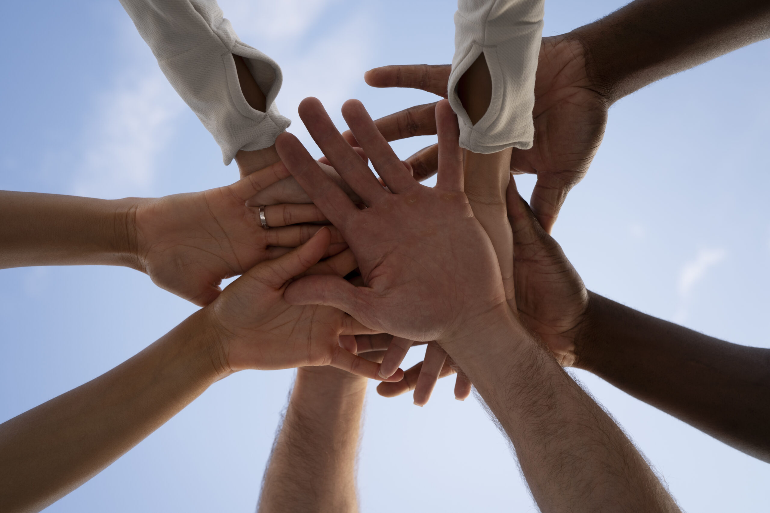Un groupe de mains de différentes ethnies se superpose, unies vers le haut, sur fond de ciel bleu. Certaines mains portent des bagues ou des bracelets, illustrant une collaboration et une connexion entre les individus.