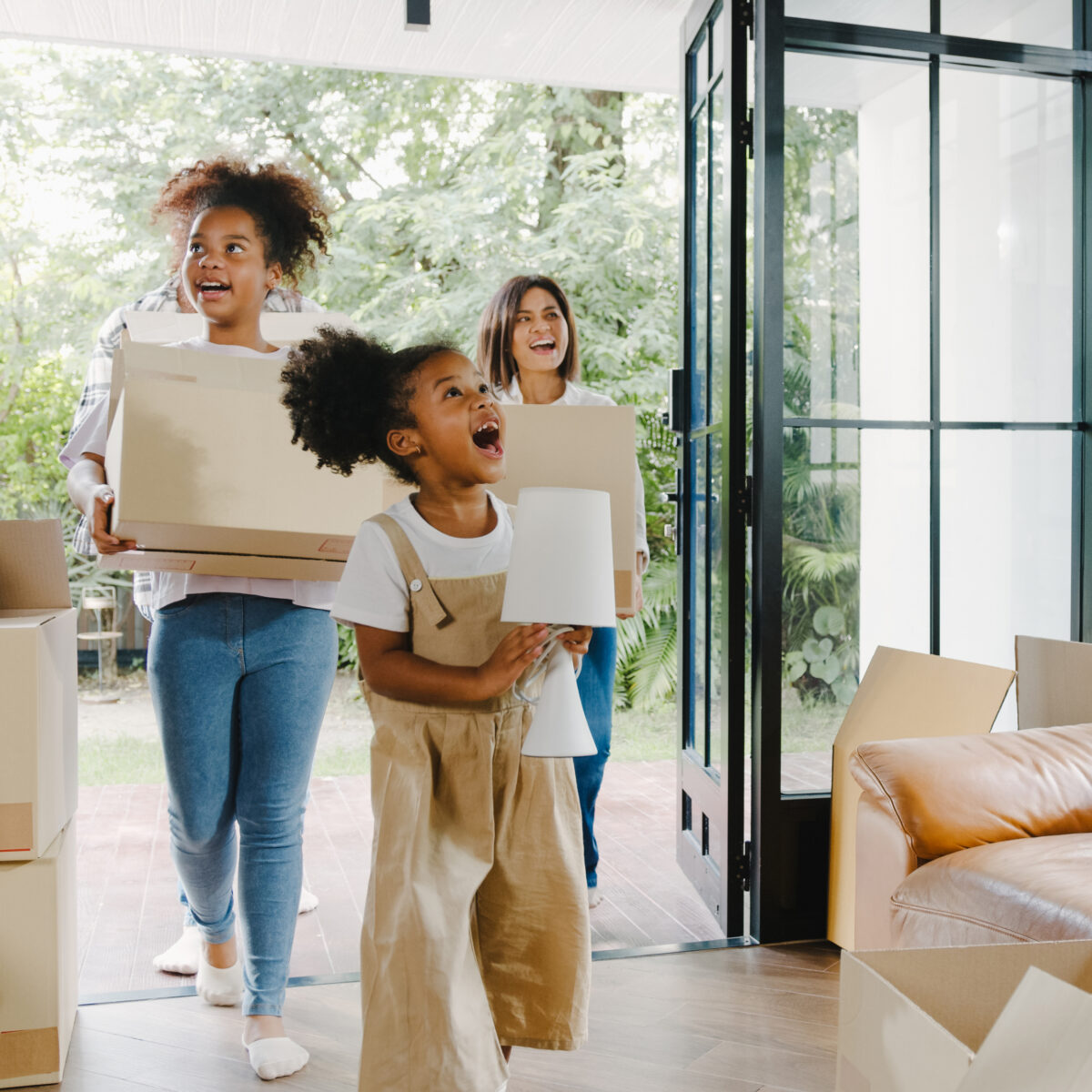Trois personnes, dont deux enfants, entrent dans une maison en portant des cartons. L'une des enfants, joyeuse, tient une lampe, tandis qu'une adulte souriante transporte un carton. L'intérieur est lumineux et décoré de plantes, avec des cartons de déménagement éparpillés autour d'un canapé en cuir.