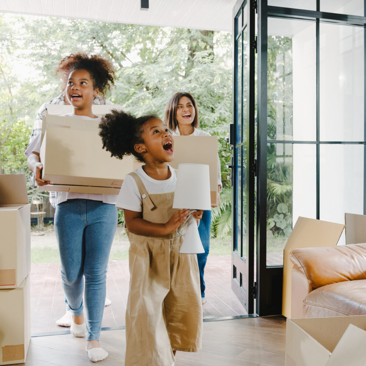 Trois personnes, dont deux enfants, entrent dans une maison en portant des cartons. L'une des enfants, joyeuse, tient une lampe, tandis qu'une adulte souriante transporte un carton. L'intérieur est lumineux et décoré de plantes, avec des cartons de déménagement éparpillés autour d'un canapé en cuir.