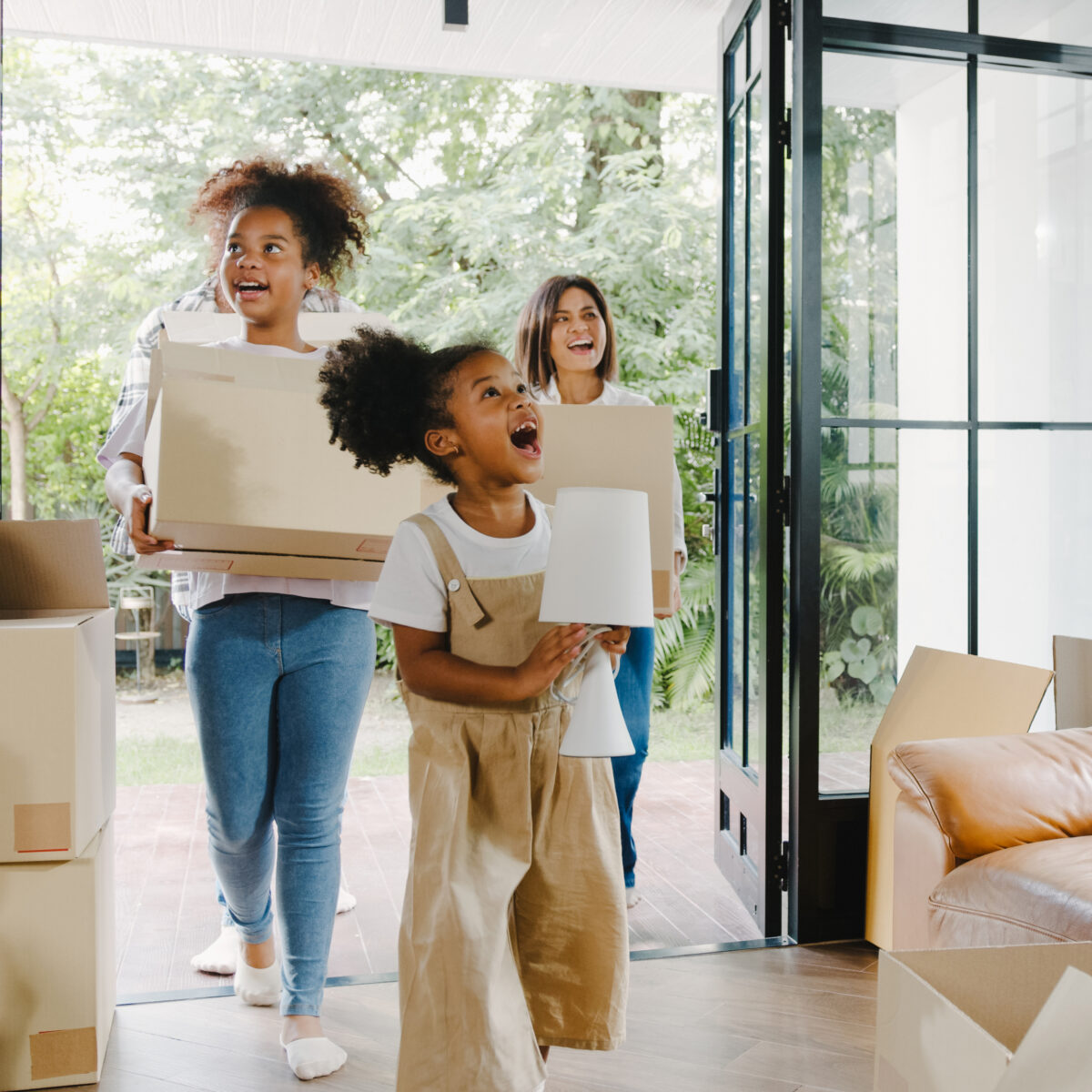 Trois personnes, dont deux enfants, entrent dans une maison en portant des cartons. L'une des enfants, joyeuse, tient une lampe, tandis qu'une adulte souriante transporte un carton. L'intérieur est lumineux et décoré de plantes, avec des cartons de déménagement éparpillés autour d'un canapé en cuir.