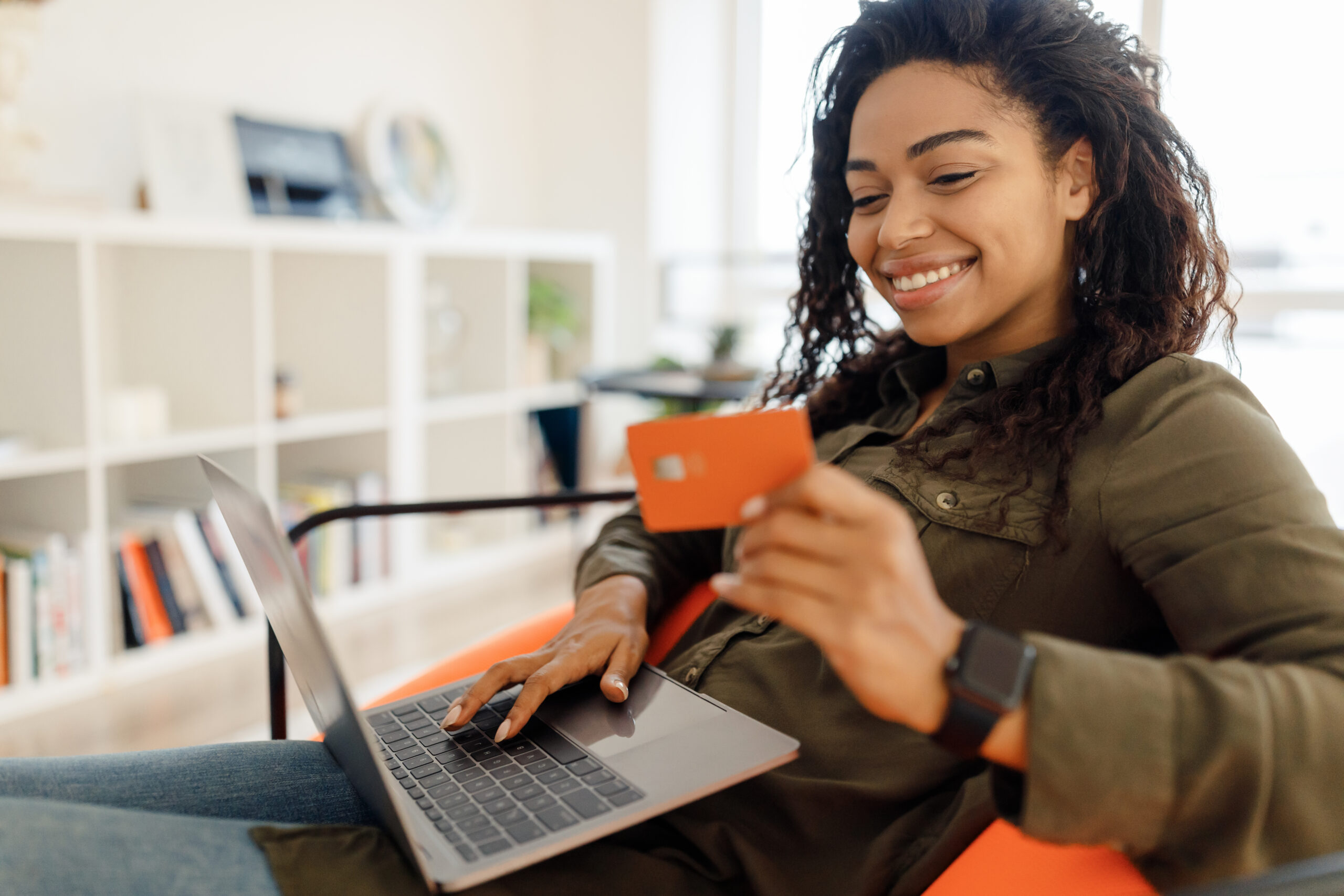 Concept d'achat en ligne. Portrait d'une acheteuse noire souriante utilisant un ordinateur portable tenant une carte de crédit de débit achetant des choses sur la boutique en ligne assise sur une chaise pouf à la maison.