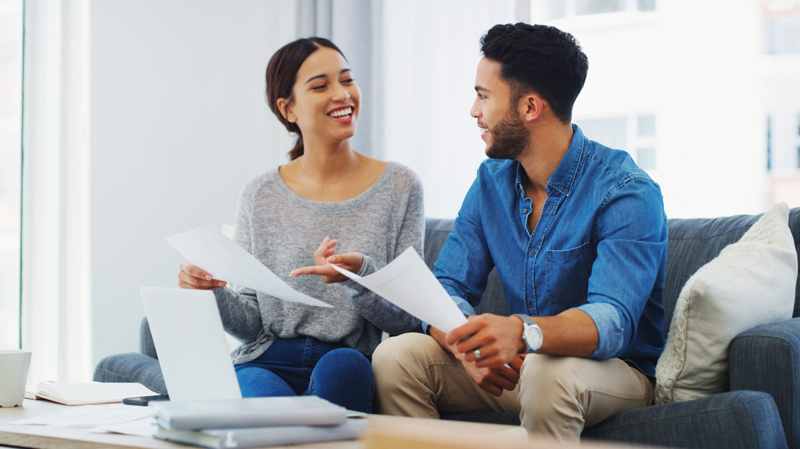 Un jeune couple assis dans son canapé, échange sur des documents administratifs qu'ils tiennent en main.