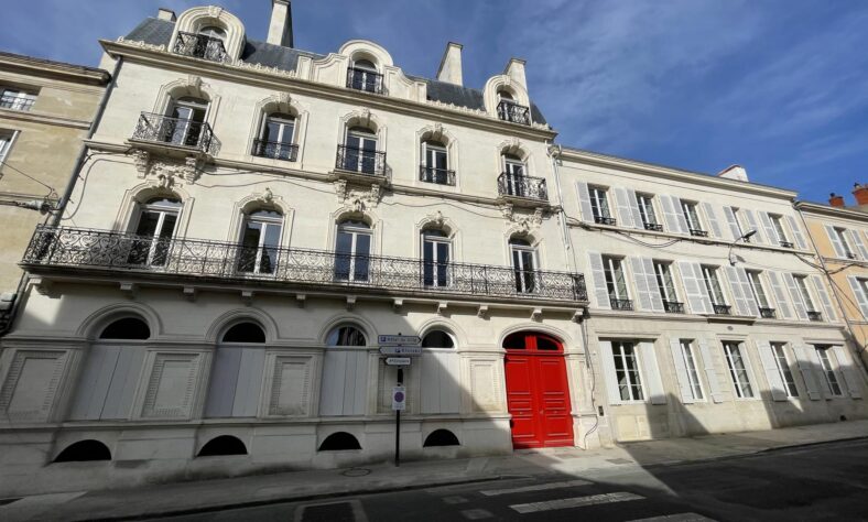 Image d'un bâtiment historique en facade blanche avec des balcons en fer forgé. La porte d'entrée est rouge, et l'architecture présente des détails élégants. En bas, on peut voir des panneaux de bois fermant les fenêtres de certains niveaux. Des panneaux de signalisation sont présents au coin de la rue. Le ciel est clair, suggérant une belle journée.
