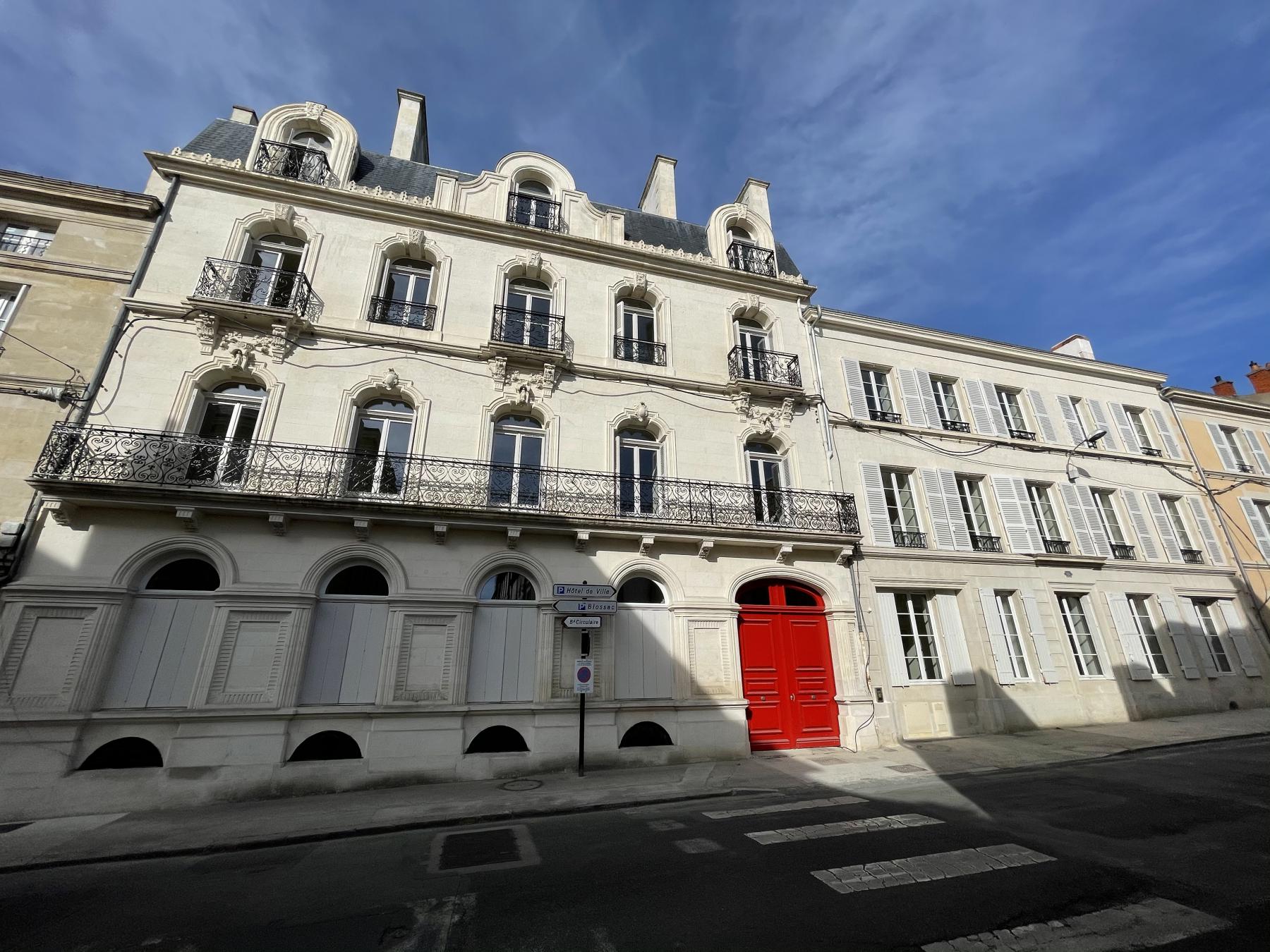Image d'un bâtiment historique en facade blanche avec des balcons en fer forgé. La porte d'entrée est rouge, et l'architecture présente des détails élégants. En bas, on peut voir des panneaux de bois fermant les fenêtres de certains niveaux. Des panneaux de signalisation sont présents au coin de la rue. Le ciel est clair, suggérant une belle journée.