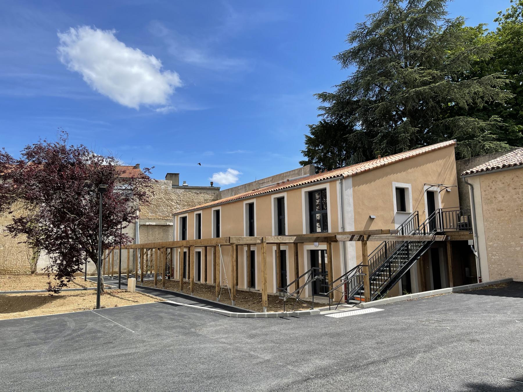 Une vue d'un bâtiment moderne avec plusieurs étages, comportant de grandes fenêtres. À l'avant, un chemin en bois mène à un escalier menant à l'entrée. On aperçoit un jardin avec un arbre rouge et un ciel bleu, parsemé de quelques nuages.