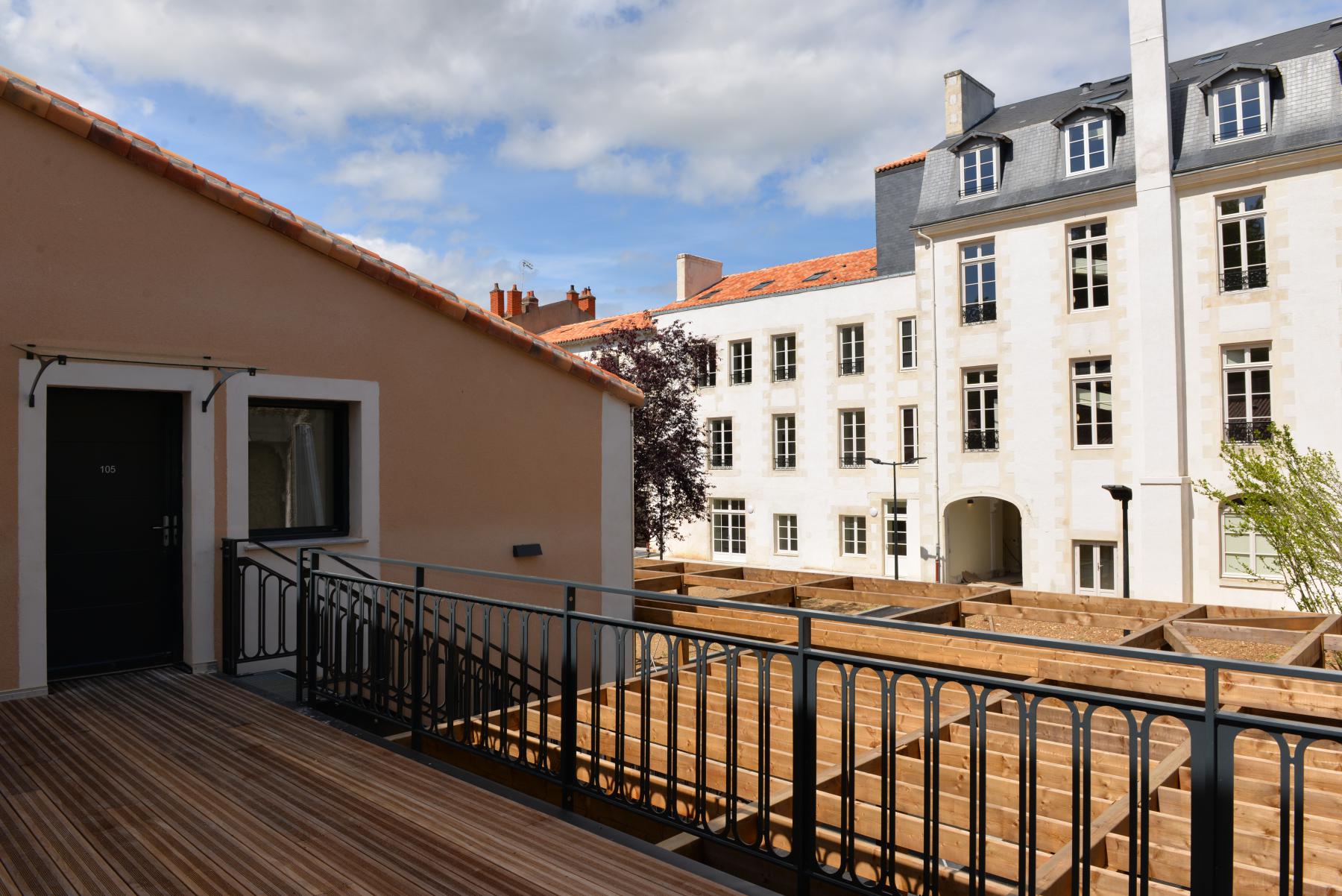 Une vue depuis un balcon, montrant un toit en tuiles et un mur beige. À l'arrière-plan, un bâtiment en pierre blanche avec de grandes fenêtres. Un espace extérieur en bois brut est visible en bas, entouré de quelques arbres. Le ciel est partiellement nuageux.