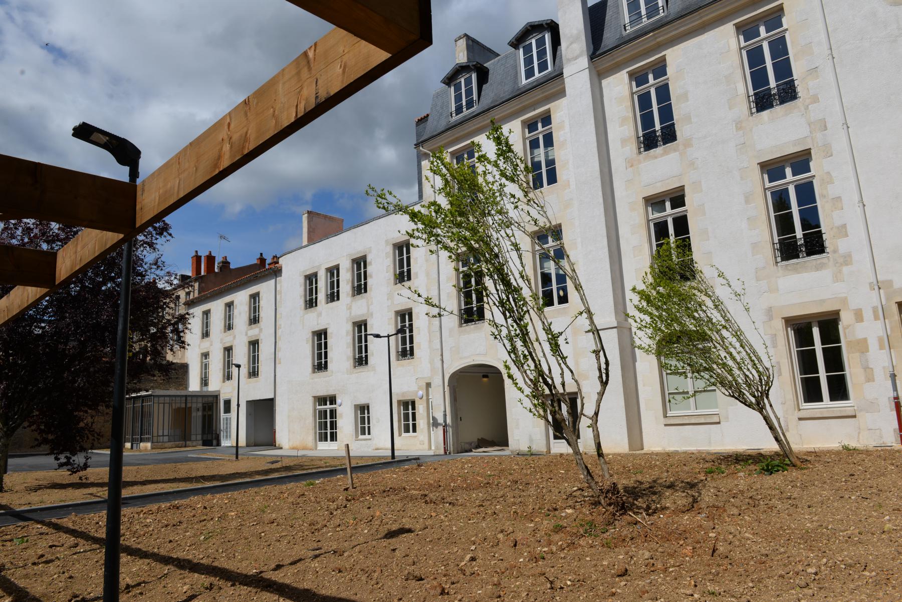 Un bâtiment de style contemporain, en pierres claires, avec plusieurs fenêtres à petits carreaux et un toit à plusieurs pentes. L'espace extérieur est en cours d'aménagement, avec quelques jeunes arbres et un sol nu. À gauche, une structure en bois est visible, ainsi qu'un éclairage. Le ciel est partiellement nuageux.