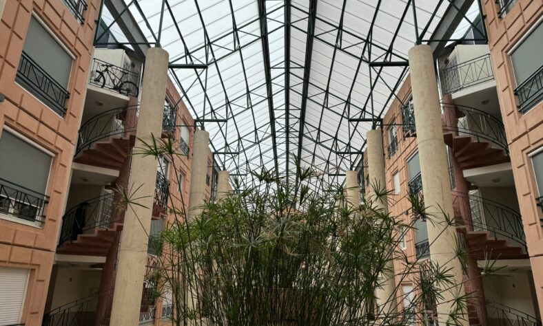 L'image montre l'intérieur d'un bâtiment avec un grand plafond en verre. Il y a plusieurs étages avec des escaliers courbes et des balcons de chaque côté. De hautes plantes vertes sont au centre, s'étendant vers le plafond.