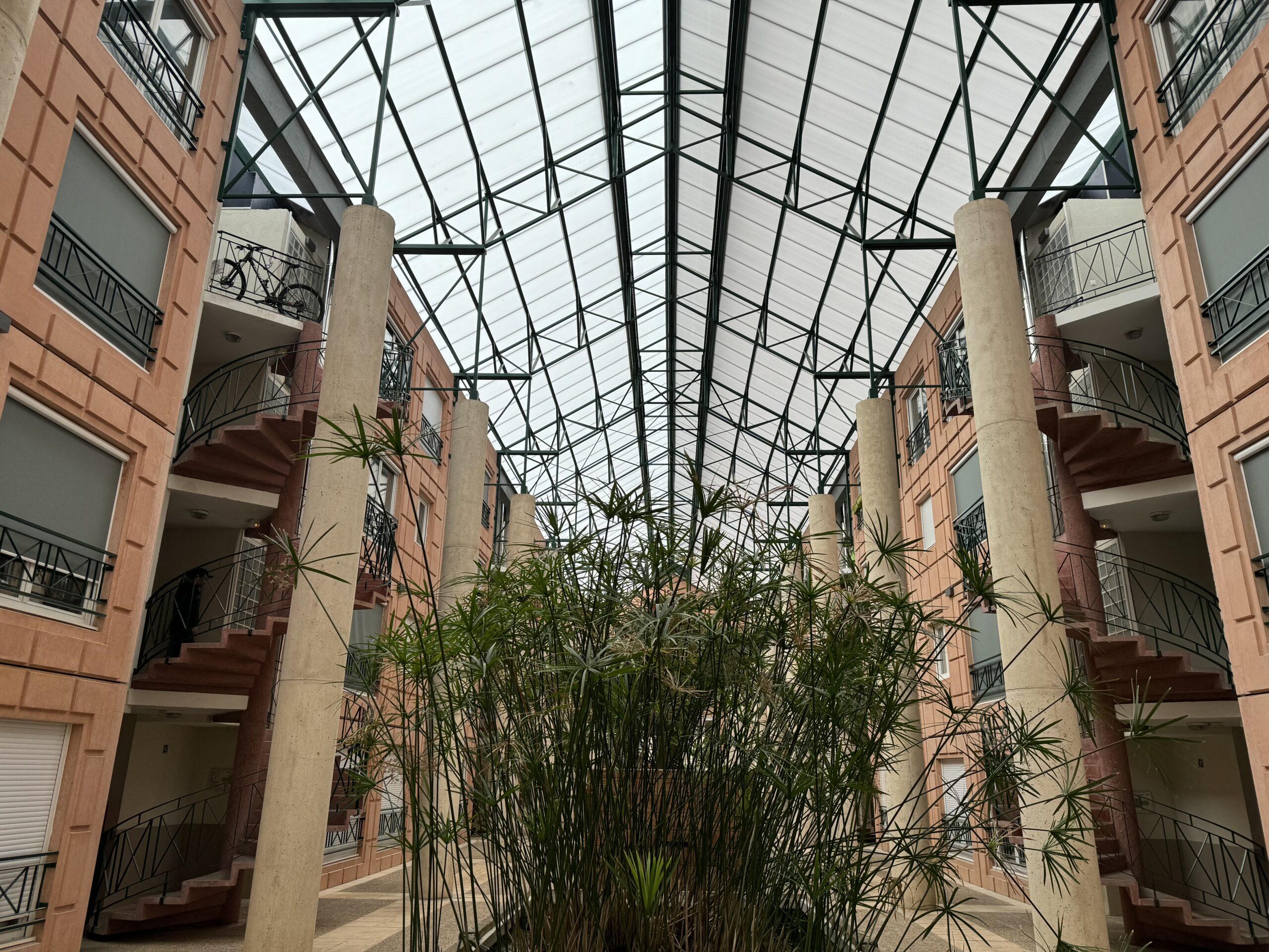 L'image montre l'intérieur d'un bâtiment avec un grand plafond en verre. Il y a plusieurs étages avec des escaliers courbes et des balcons de chaque côté. De hautes plantes vertes sont au centre, s'étendant vers le plafond.