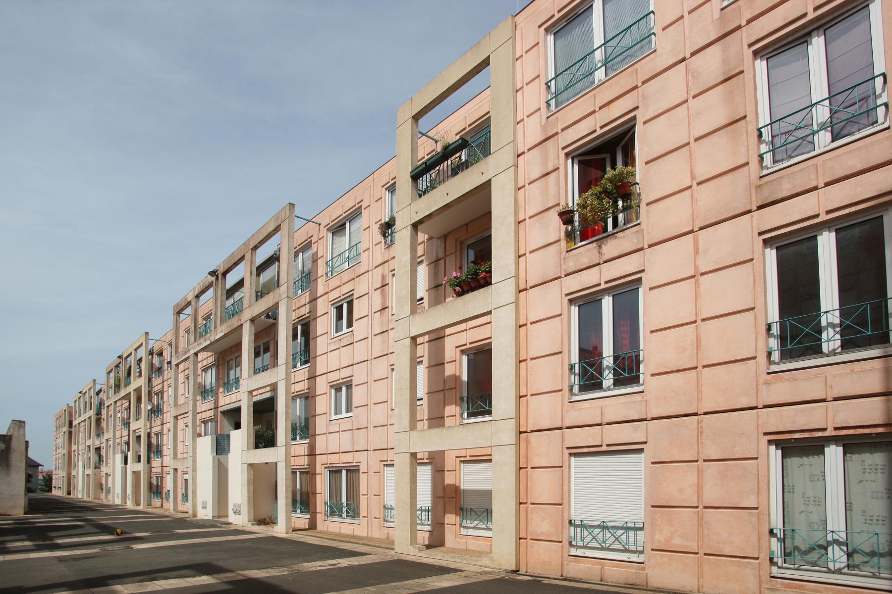 Une rangée d'immeubles d'appartements beiges et géométriques avec de nombreux balcons et fenêtres sous un ciel clair. Certains balcons sont plantés de plantes. La rue à côté des immeubles semble vide, créant une atmosphère sereine et spacieuse.