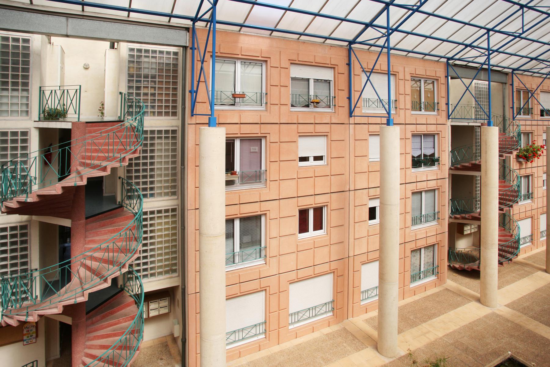 On voit ici un immeuble d'appartements moderne avec une façade orange pâle. Il est doté d'escaliers en colimaçon, de grandes fenêtres carrées et de poutres métalliques bleues visibles sous un toit translucide. Il y a des balcons avec des plantes sur le côté droit.