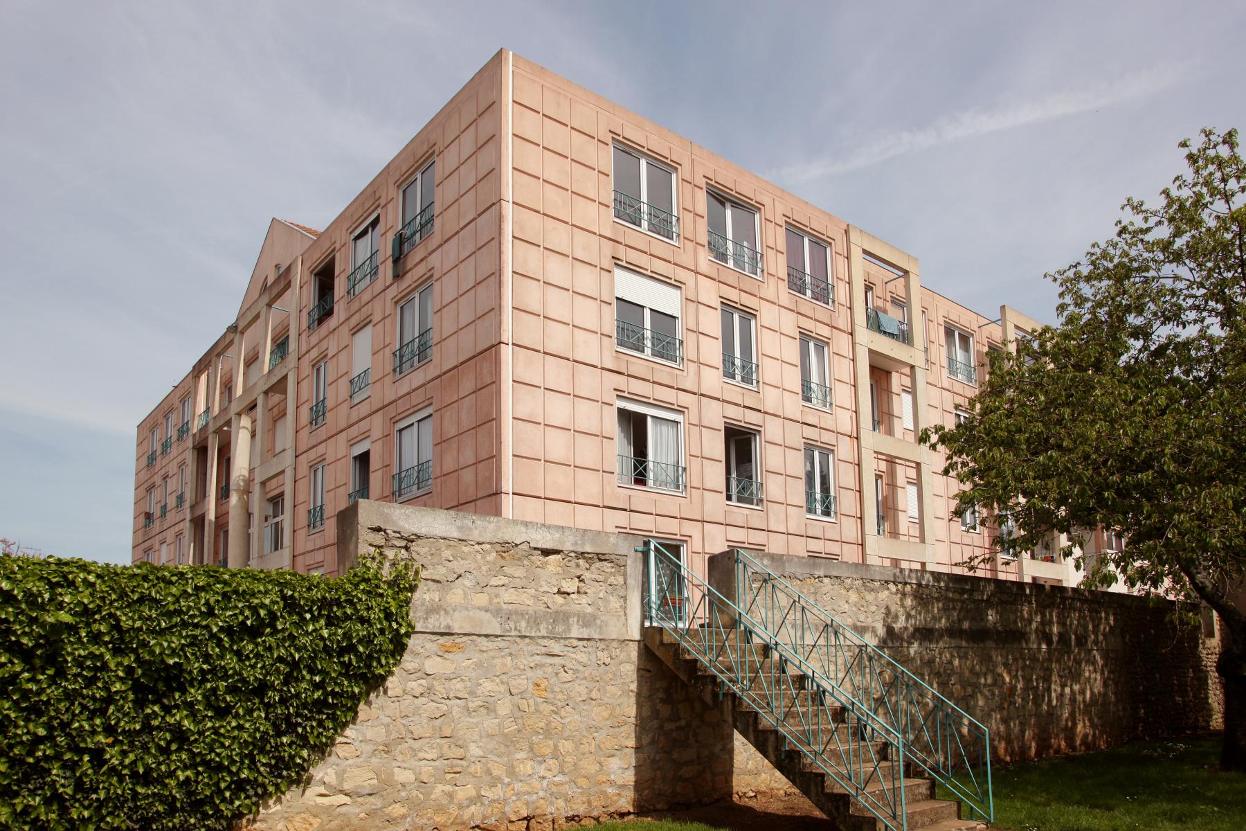 Derrière un mur de pierre se dresse un immeuble d'appartements beige rosé avec de multiples fenêtres. Un escalier en métal mène au bâtiment. Des arbres et de la verdure entourent la scène sous un ciel partiellement nuageux.