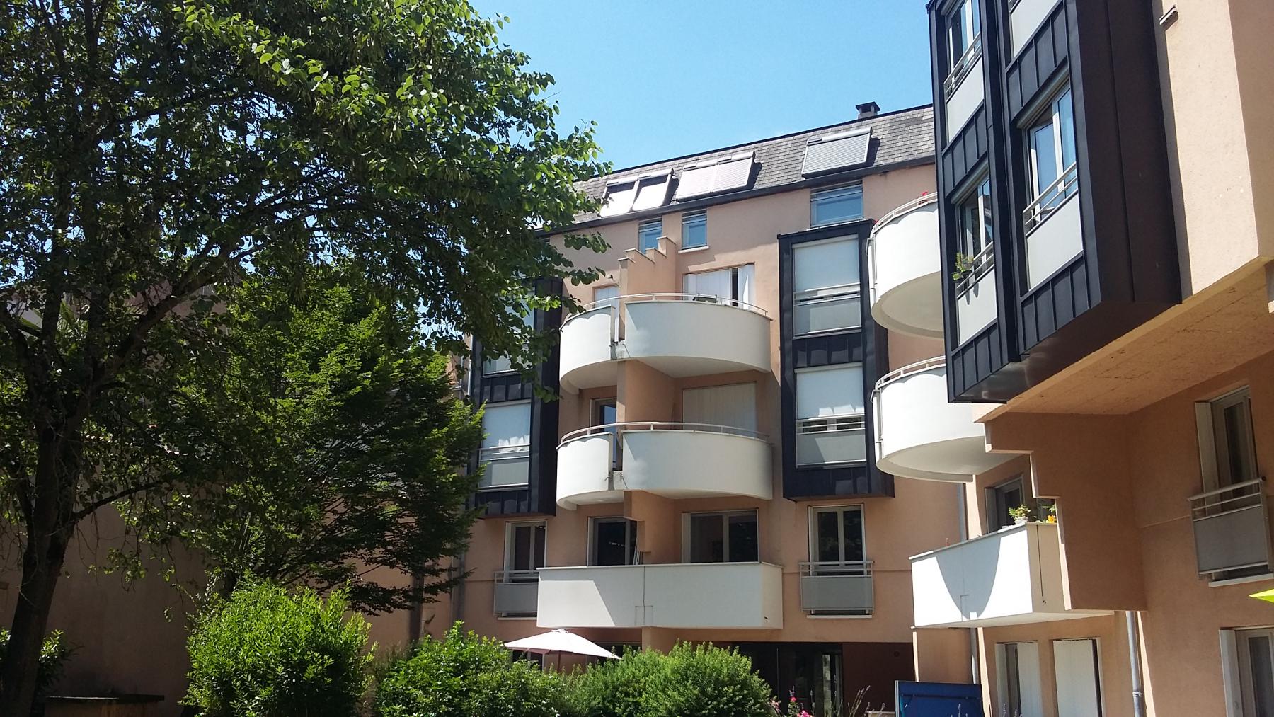 Immeuble d'appartements moderne avec balcons incurvés et grandes fenêtres. Il est entouré d'une végétation luxuriante et d'arbres, sous un ciel bleu clair. Le bâtiment présente un mélange de tons pêche clair et gris foncé.