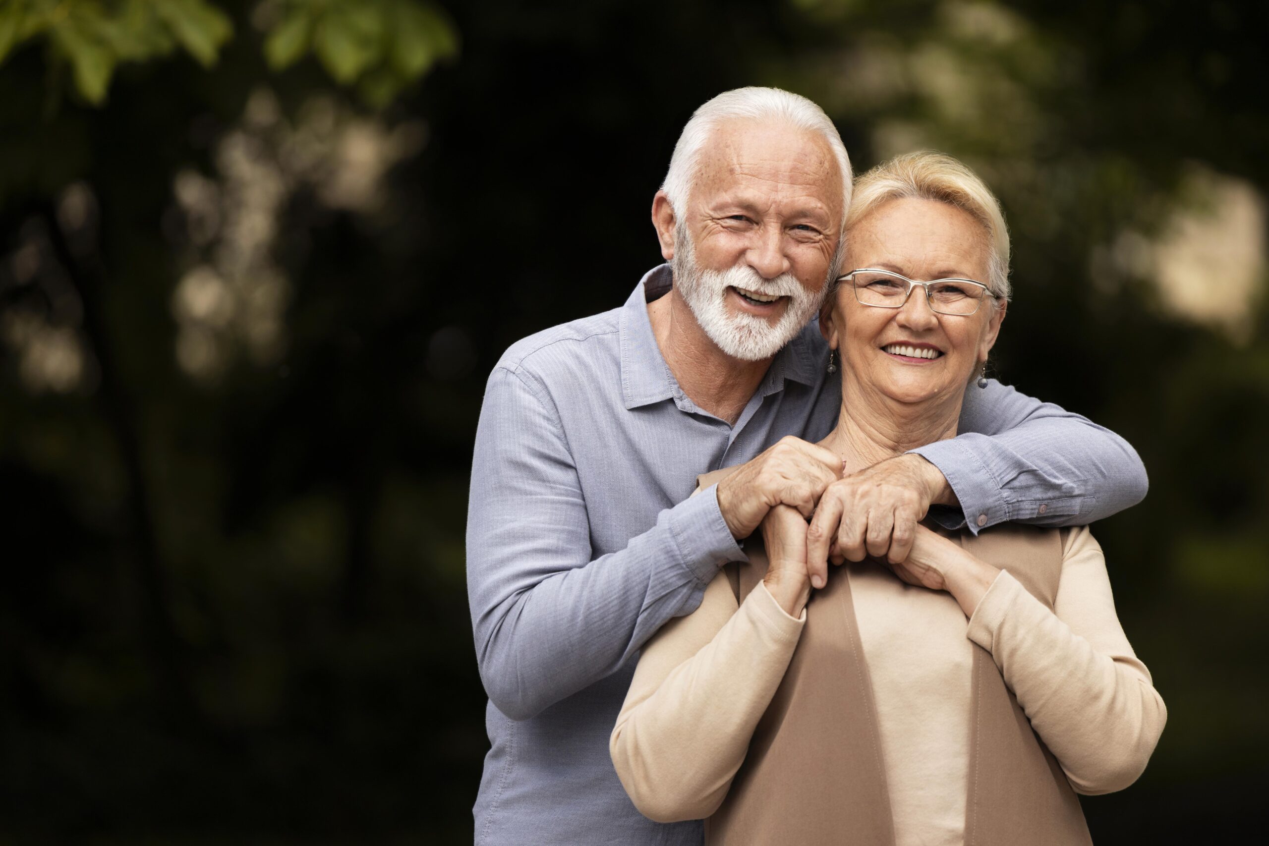 Un couple de personnes âgées, l'homme enlaçant la femme par derrière. Ils semblent tous deux joyeux, l'homme portant une barbe blanche et la femme portant des lunettes. Ils sont habillés de manière décontractée, l'homme portant une chemise bleu clair et la femme un haut beige. L'arrière-plan est flou, mettant en valeur le couple, mais laisse entrevoir un cadre naturel avec de la verdure.